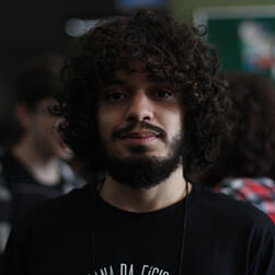 Um círculo com uma fotografia colorida, ambiente interno. Uma foto do Pedro até o começo dos ombros de camiseta preta. Dá para ver pessoas no fundo. Pedro é branco, tem o cabelo cacheado castanho em torno do rosto, barba e bigode. Está com um semissorriso.