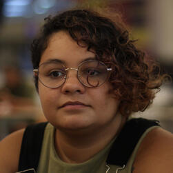 fotografia colorida, ambiente interno. Uma foto da Luisa dos ombros para cima. Da para ver a blusa verde oliva e as alças de um macacão preto. Ela tem a pele bege e a cara redonda. O fundo da foto é um lugar público e está desfocado. Ela está de óculos de